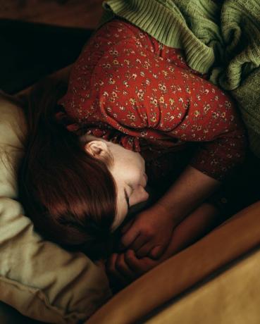 صورة لامرأة ذات شعر أحمر مغطاة ببطانية خضراء مستلقية على الأريكة الصفراء وتنام بمزاج الخريف أو الشتاء وجو دافئ ومريح يشرب قهوة الصباح