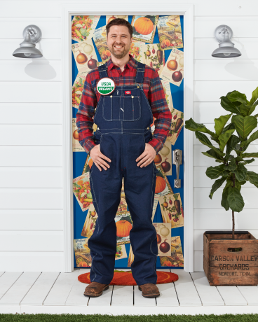 رجل يرتدي زي المزارعين مع وزرة وقميص منقوش وزر عضوي من وزارة الزراعة الأمريكية يقف أمام الباب مزينًا بالهالوين