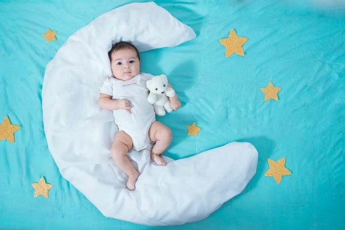 طفلة لاتينية جميلة ، عمرها شهرين ، مستلقية على ورقة بيضاء على شكل قمر مع نجوم صفراء على كل جانب وصفيحة زرقاء تحتها كلها