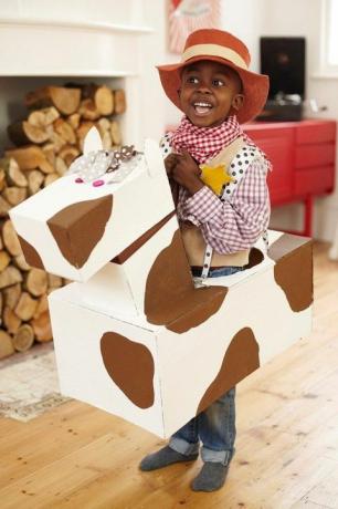 صبي صغير يرتدي زي راعي البقر مع قبعة رعاة البقر وقميص منقوش وباندانا مع حصان من الورق المقوى حول خصره