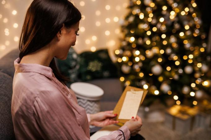 شابة سعيدة تجلس على الأريكة بجوار شجرة عيد الميلاد المتلألئة، وتفتح وتقرأ بطاقة عيد الميلاد التي حصلت عليها مع هديتها