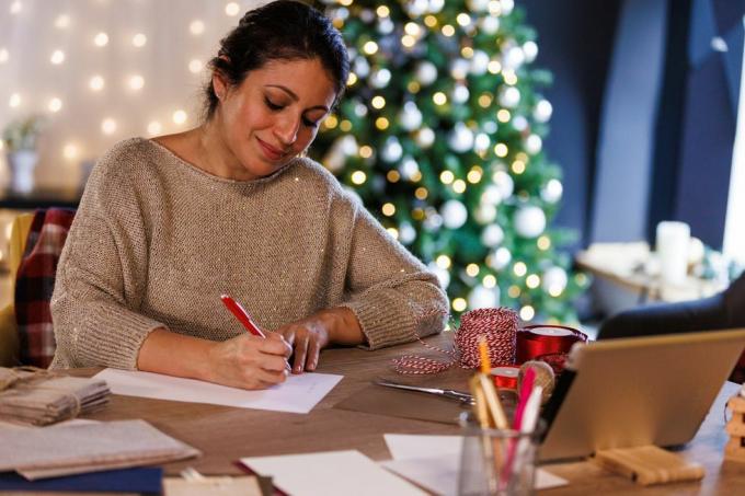 صورة لامرأة سعيدة في منتصف العمر تجلس على الطاولة أمام شجرة عيد الميلاد المتلألئة، وتبتسم أثناء كتابة بطاقات عيد الميلاد والرسائل للأحباء