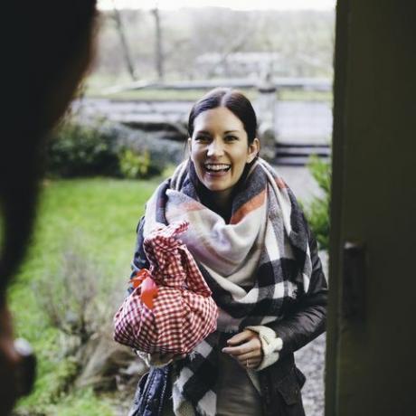 امرأة تبتسم وهي تقف على درج الباب تزور صديقها وهي ملفوفة بملابس دافئة وتحمل هدية ملفوفة بقطعة قماش حمراء وبيضاء مربعة