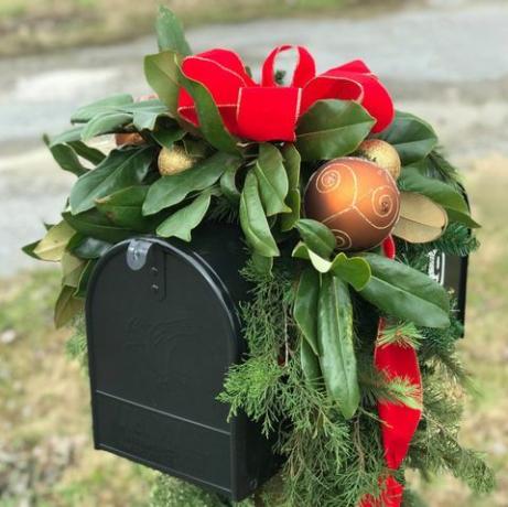 صندوق بريد أسود مزين بالكرات الذهبية للكريسماس وشريط أحمر والكثير من المساحات الخضراء بما في ذلك أوراق الماغنوليا والأرز