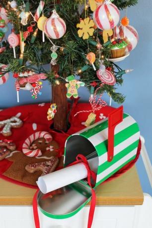 صندوق بريد صغير مخطط باللونين الأبيض والأخضر مع ورق ملفوف مربوط بشريط أحمر فيه ، ويجلس تحت شجرة عيد الميلاد الصغيرة ، على تنورة شجرة عيد الميلاد الحمراء