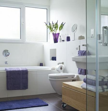 حمام مضاء بشكل مشرق مع منشفة أرجوانية على جانب الحمام ومناشف مطوية بالقرب من الدش